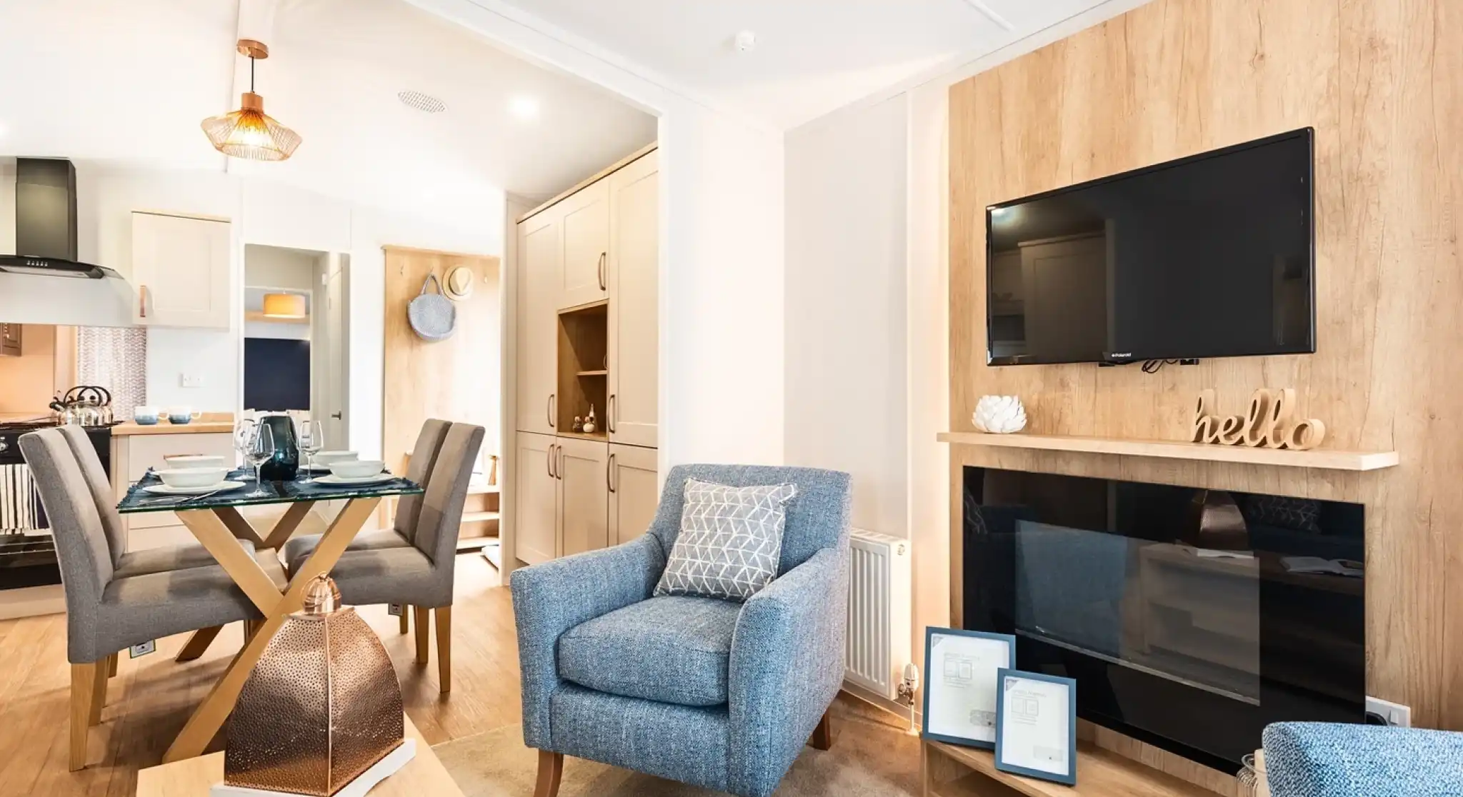 A feature wooden wall with electric fireplace and TV above, sat in a room with cosy armchairs and a dining room and kitchen in the background.