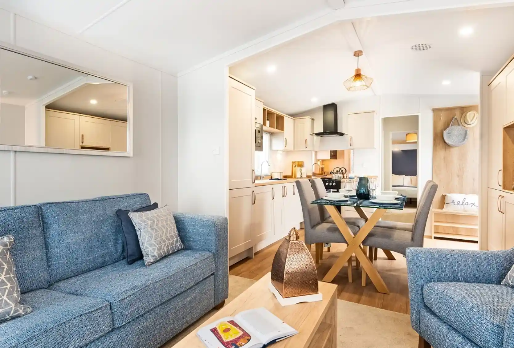 A light and airy lounge, with kitchen and dining area. There is a blue comfortable sofa with coffee table and armchair in the foreground. A glass dining table with four chairs and cream kitchen cabinets sit in the background.