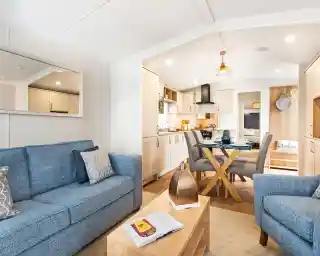 A light and airy lounge, with kitchen and dining area. There is a blue comfortable sofa with coffee table and armchair in the foreground. A glass dining table with four chairs and cream kitchen cabinets sit in the background.