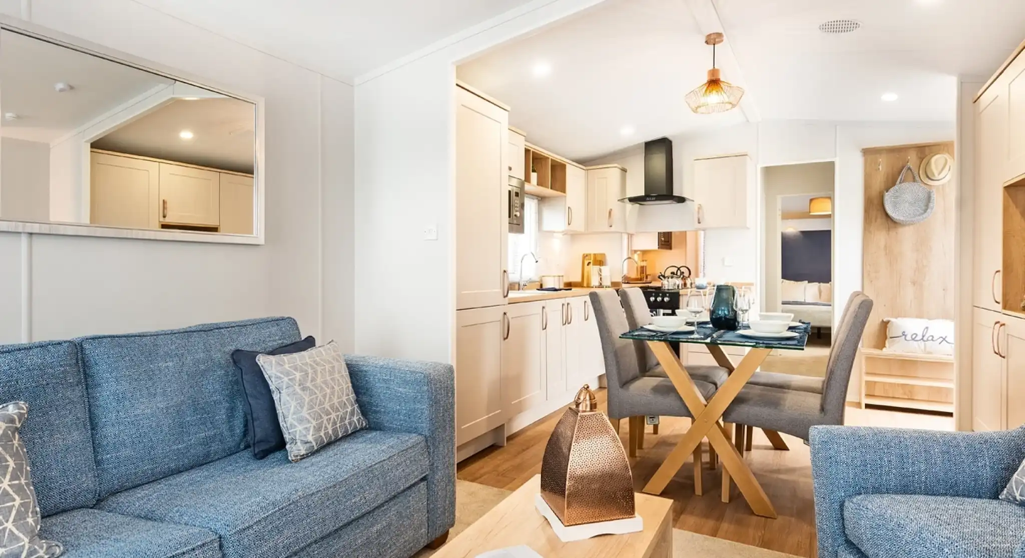 A light and airy lounge, with kitchen and dining area. There is a blue comfortable sofa with coffee table and armchair in the foreground. A glass dining table with four chairs and cream kitchen cabinets sit in the background.