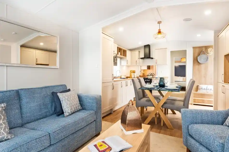 A light and airy lounge, with kitchen and dining area. There is a blue comfortable sofa with coffee table and armchair in the foreground. A glass dining table with four chairs and cream kitchen cabinets sit in the background.