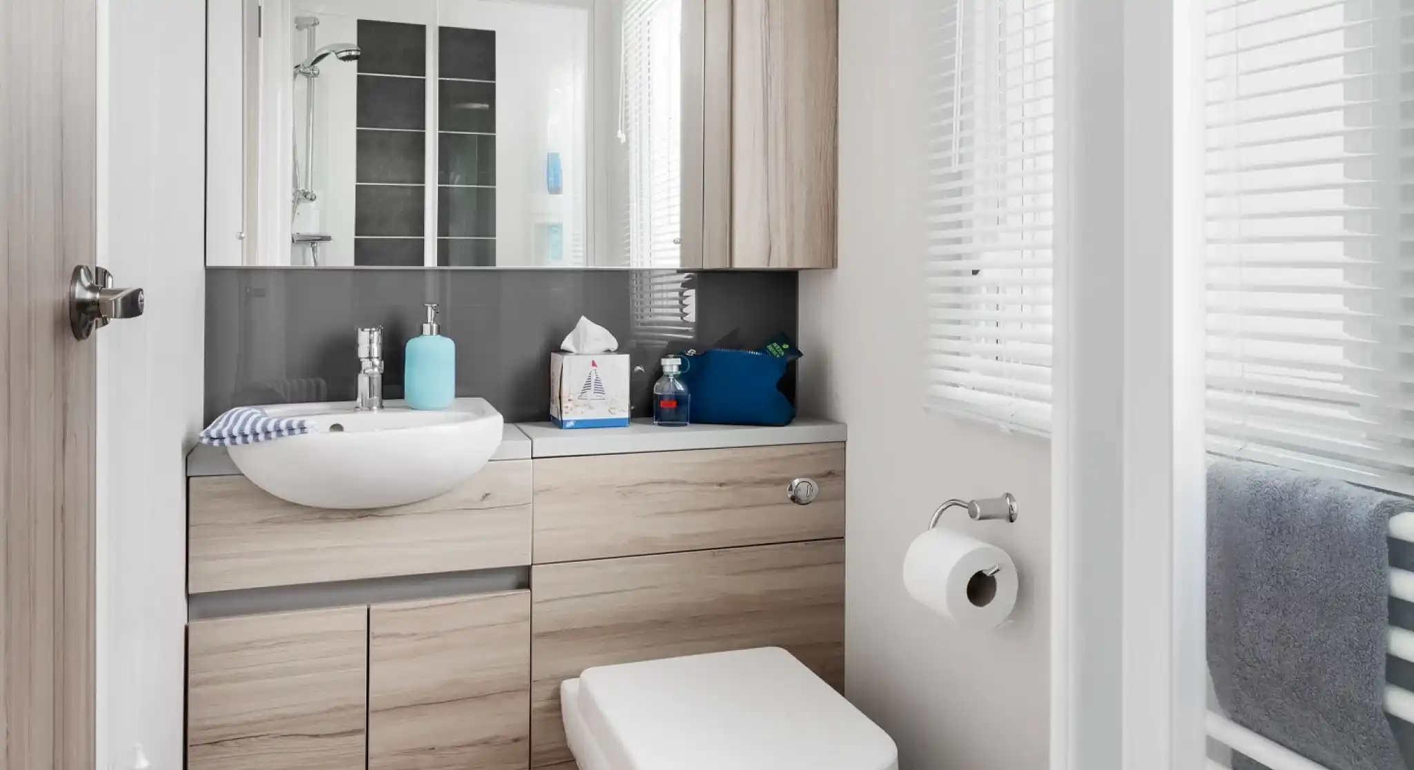 Modern bathroom with a white vessel sink, light wood vanity, and toilet next to a window with blinds.