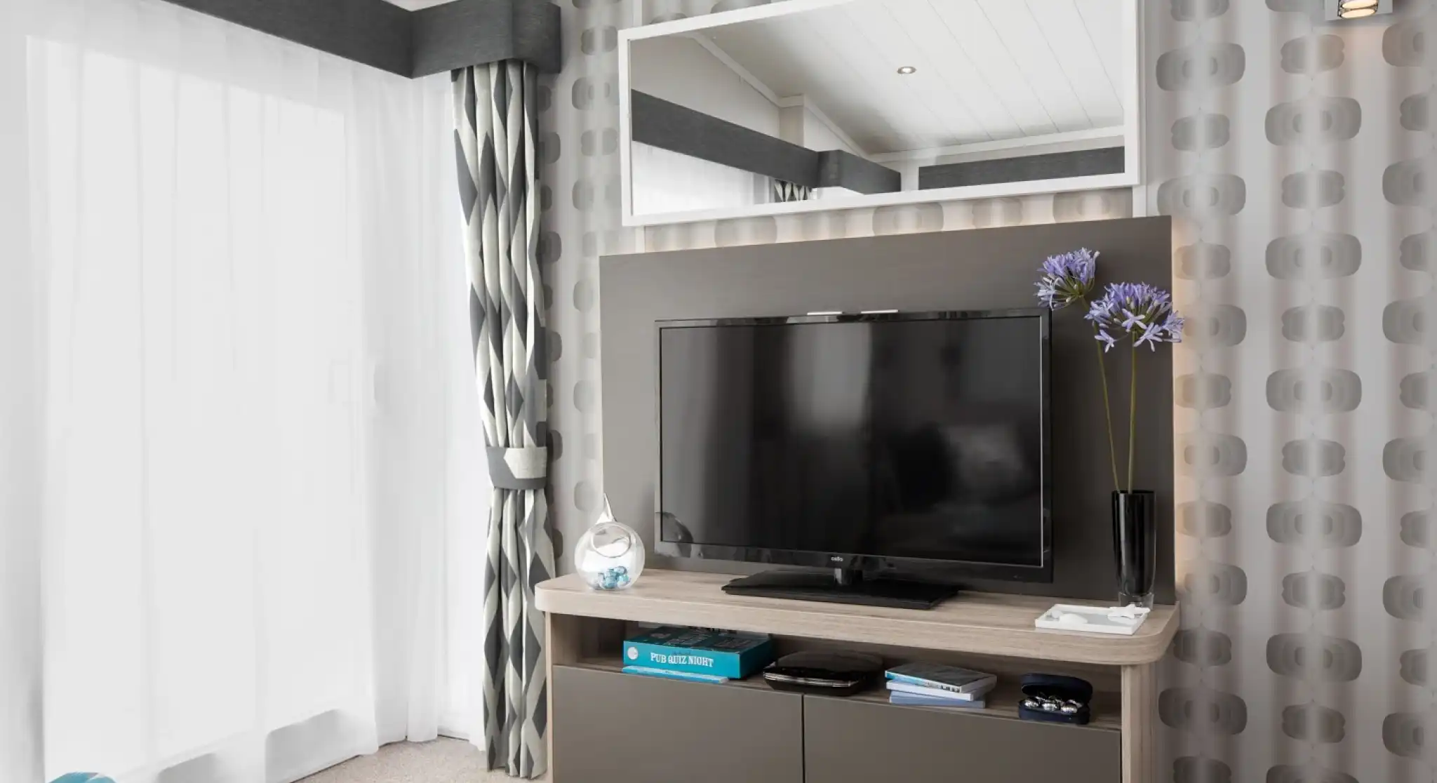 A modern living room features a wall-mounted TV on a media console, with patterned wallpaper, curtains, and a large mirror.