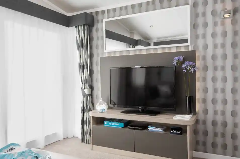A modern living room features a wall-mounted TV on a media console, with patterned wallpaper, curtains, and a large mirror.