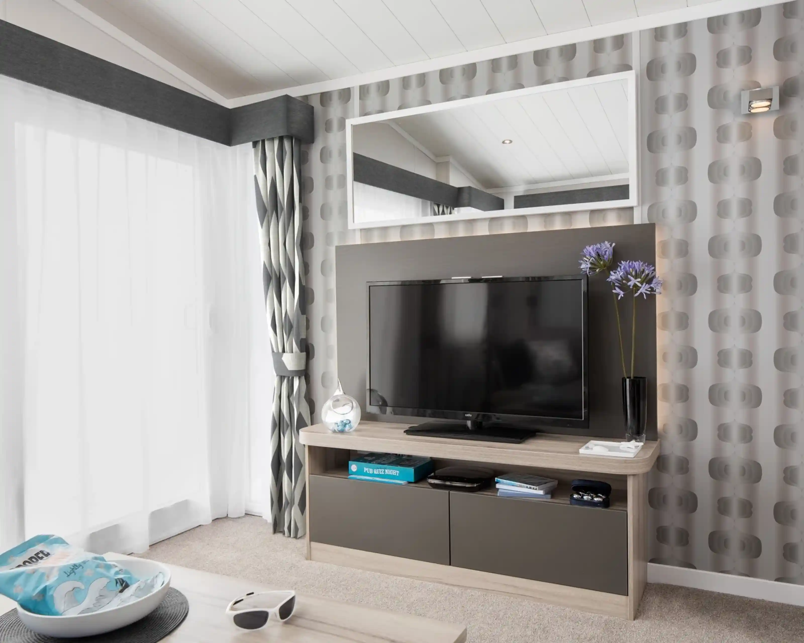 A modern living room features a wall-mounted TV on a media console, with patterned wallpaper, curtains, and a large mirror.