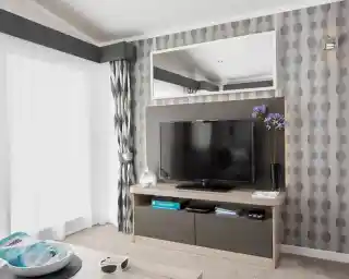 A modern living room features a wall-mounted TV on a media console, with patterned wallpaper, curtains, and a large mirror.