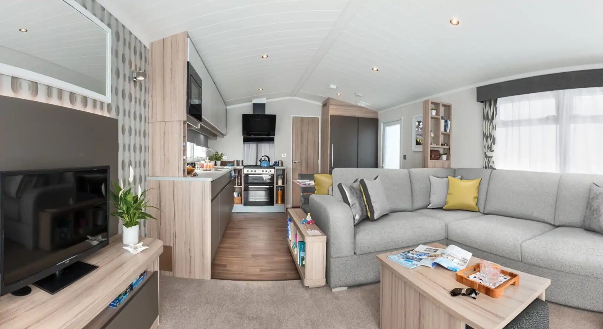 Modern caravan living area and kitchen with sectional sofa, coffee table, TV, and kitchen appliances.
