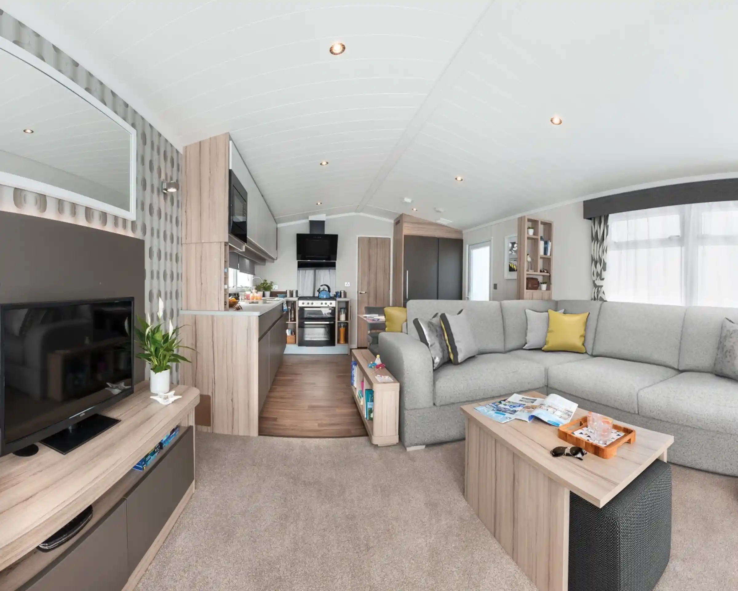 Modern caravan living area and kitchen with sectional sofa, coffee table, TV, and kitchen appliances.