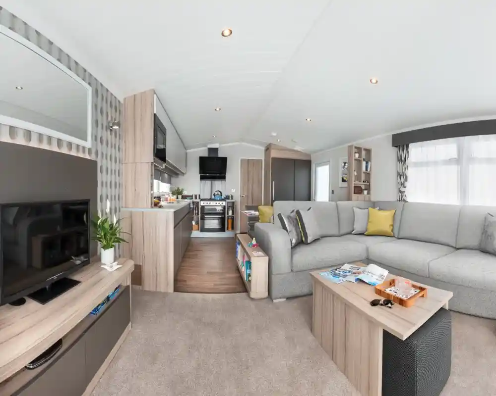 Modern caravan living area and kitchen with sectional sofa, coffee table, TV, and kitchen appliances.