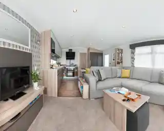 Modern caravan living area and kitchen with sectional sofa, coffee table, TV, and kitchen appliances.