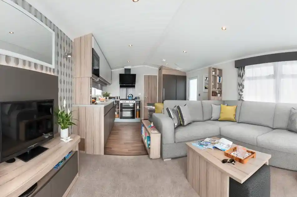 Modern caravan living area and kitchen with sectional sofa, coffee table, TV, and kitchen appliances.