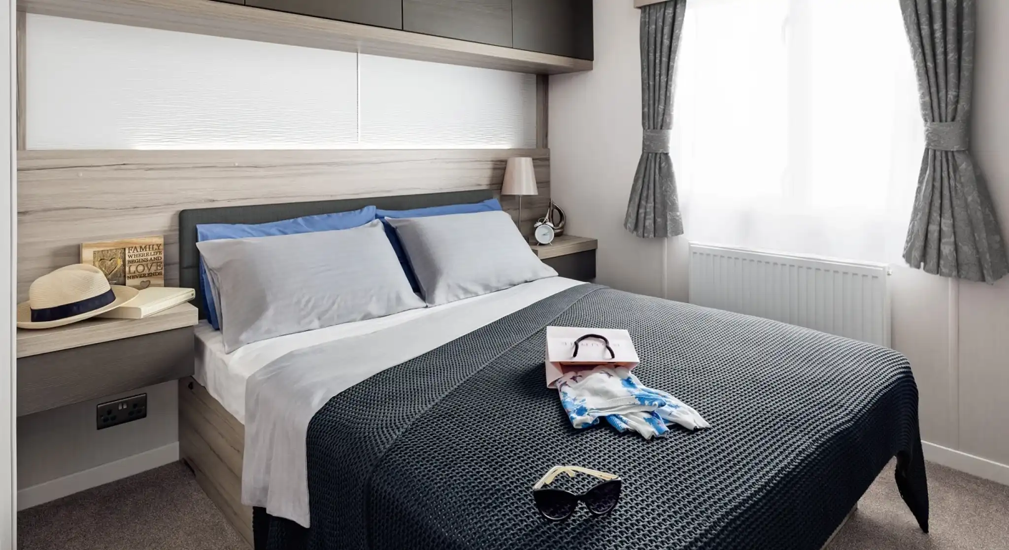 A neatly made bed with blue and grey pillows, a navy blue textured throw blanket, sunglasses, and a shopping bag with scarf.