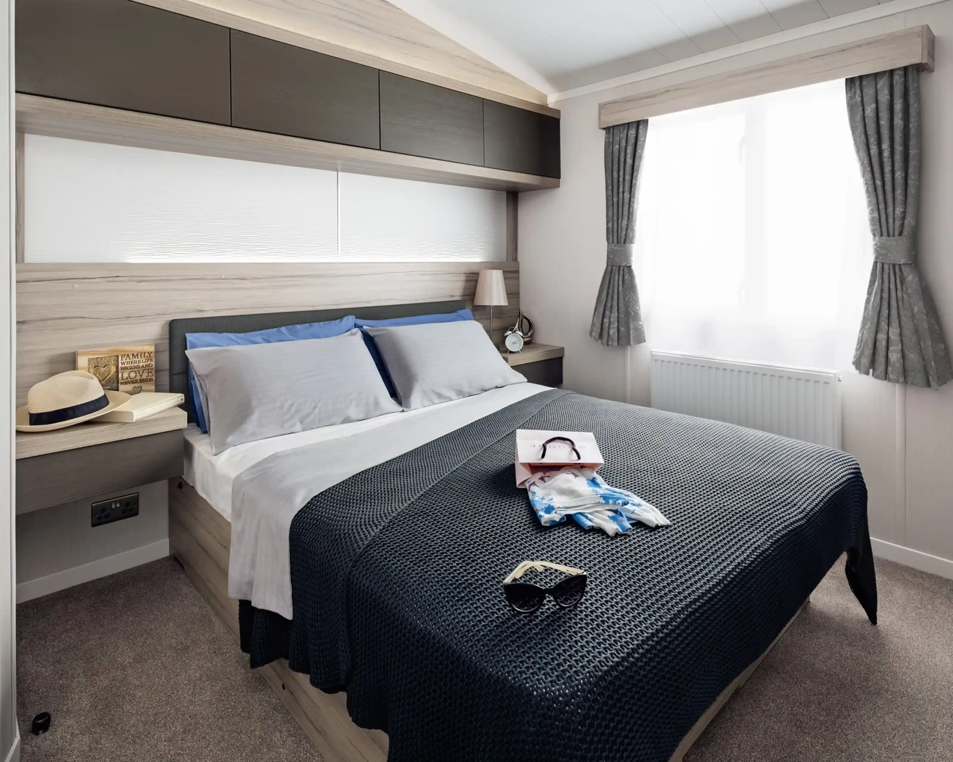A neatly made bed with blue and grey pillows, a navy blue textured throw blanket, sunglasses, and a shopping bag with scarf.