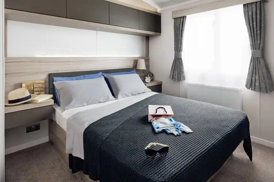 A neatly made bed with blue and grey pillows, a navy blue textured throw blanket, sunglasses, and a shopping bag with scarf.