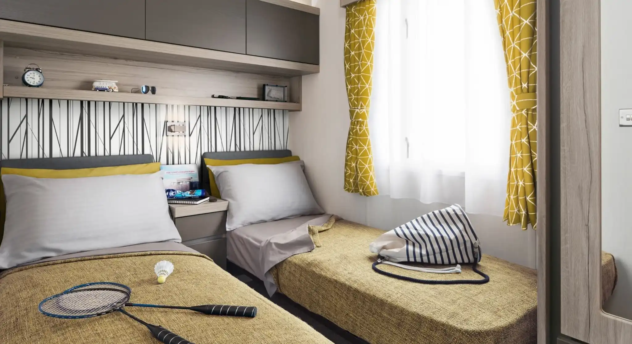 Two twin beds with yellow patterned curtains and a badminton set are in a room with a patterned wallpaper and overhead storage.