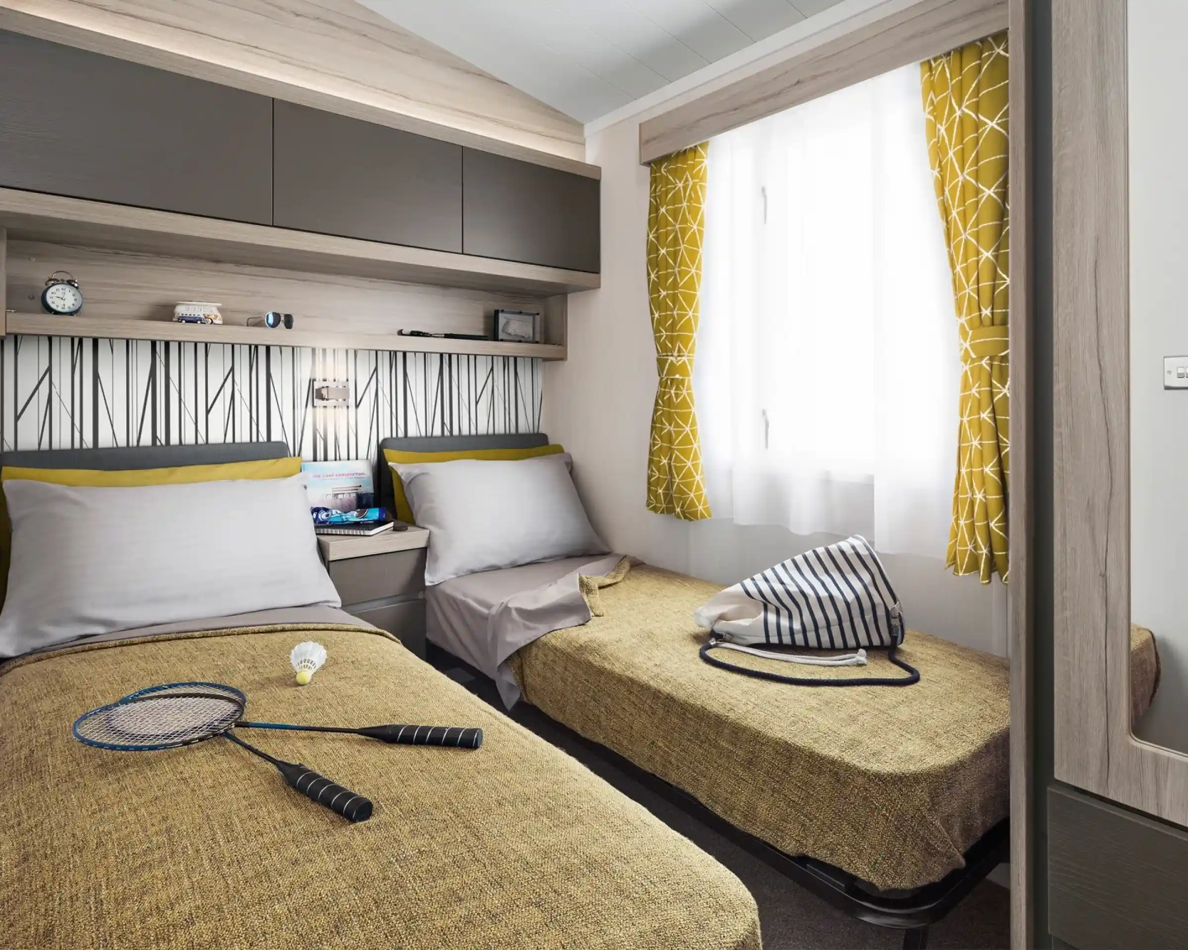 Two twin beds with yellow patterned curtains and a badminton set are in a room with a patterned wallpaper and overhead storage.