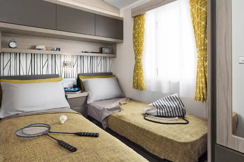 Two twin beds with yellow patterned curtains and a badminton set are in a room with a patterned wallpaper and overhead storage.