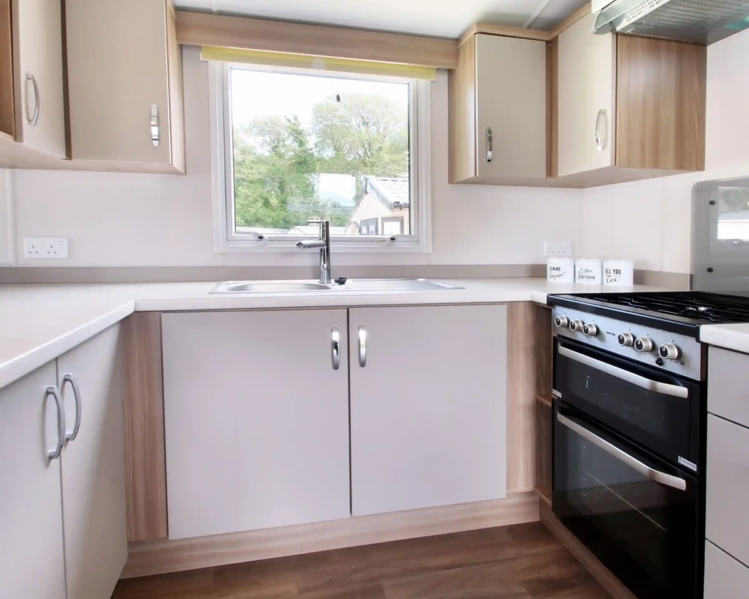 A modern kitchen featuring light-colored cabinets, a stainless steel sink, a four-burner gas stove, and a large window with a yellow blind, allowing natural light to enter.