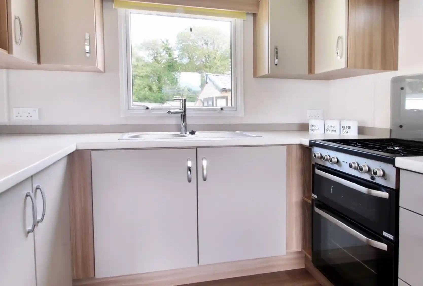 A modern kitchen featuring light-colored cabinets, a stainless steel sink, a four-burner gas stove, and a large window with a yellow blind, allowing natural light to enter.