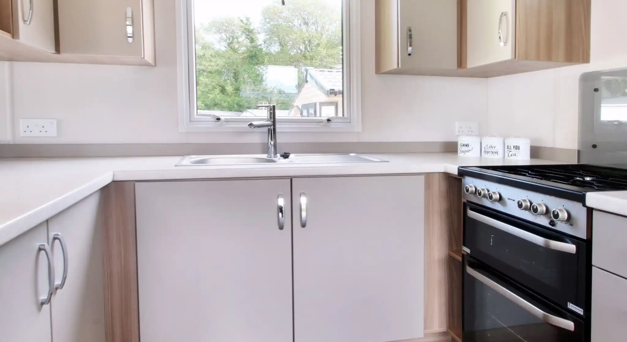 A modern kitchen featuring light-colored cabinets, a stainless steel sink, a four-burner gas stove, and a large window with a yellow blind, allowing natural light to enter.