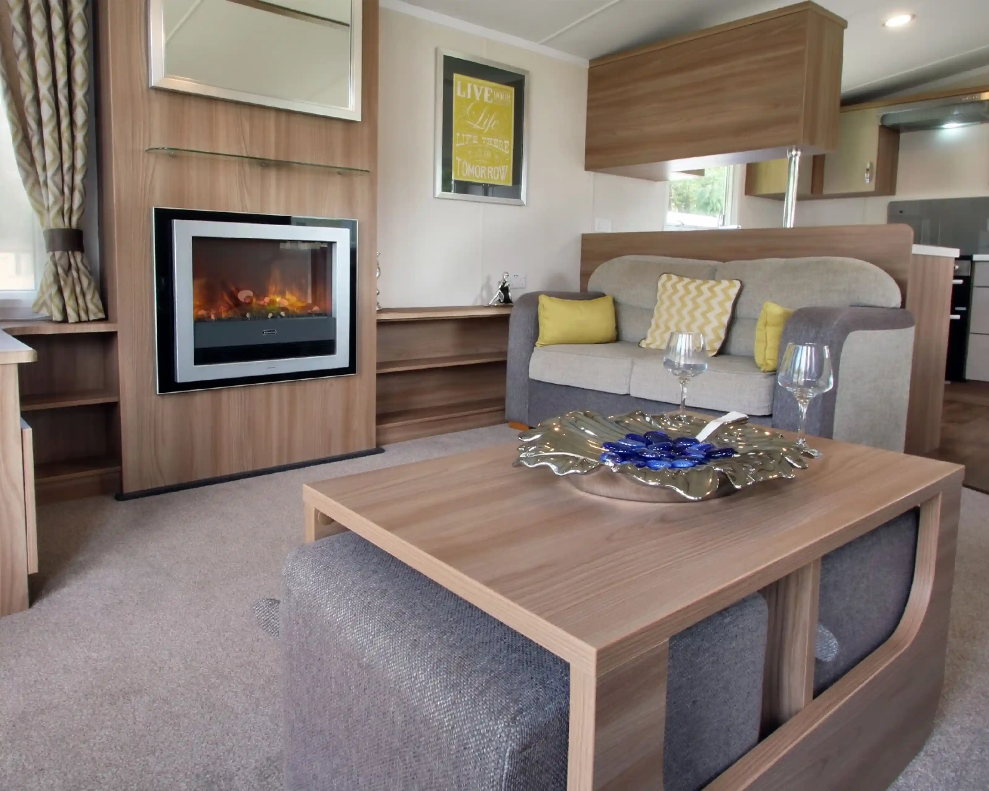 A cozy living area featuring a comfortable gray and beige sofa with decorative cushions, a modern electric fireplace, and a stylish coffee table with a decorative bowl. The room is well-lit by natural light, with curtains and wall art complementing the warm ambiance.