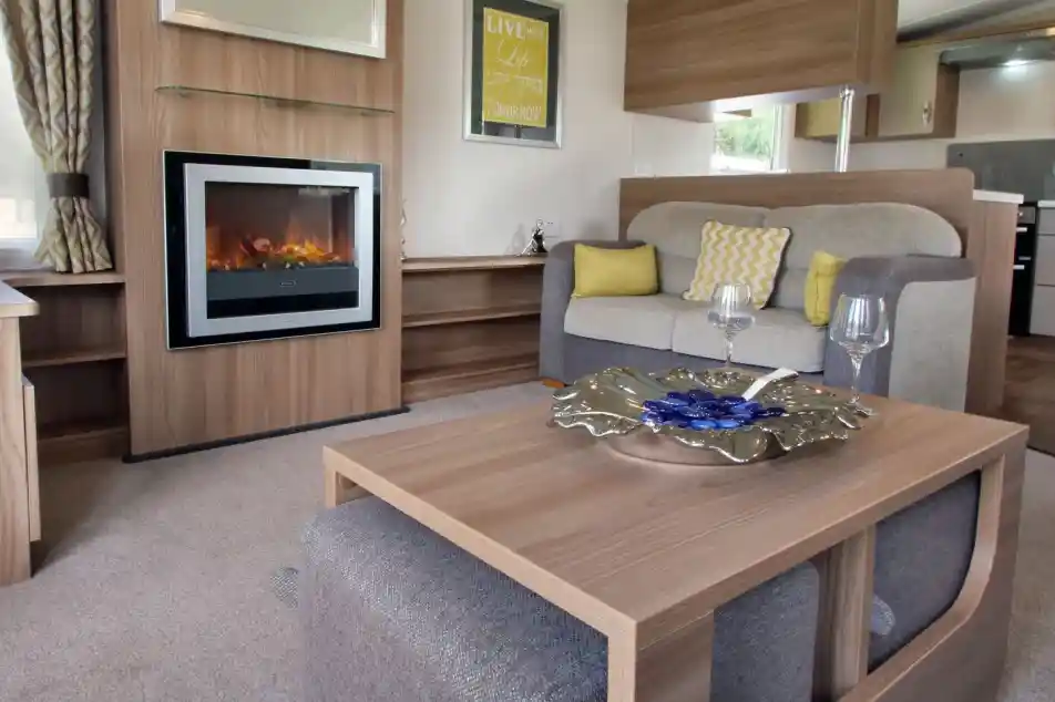 A cozy living area featuring a comfortable gray and beige sofa with decorative cushions, a modern electric fireplace, and a stylish coffee table with a decorative bowl. The room is well-lit by natural light, with curtains and wall art complementing the warm ambiance.