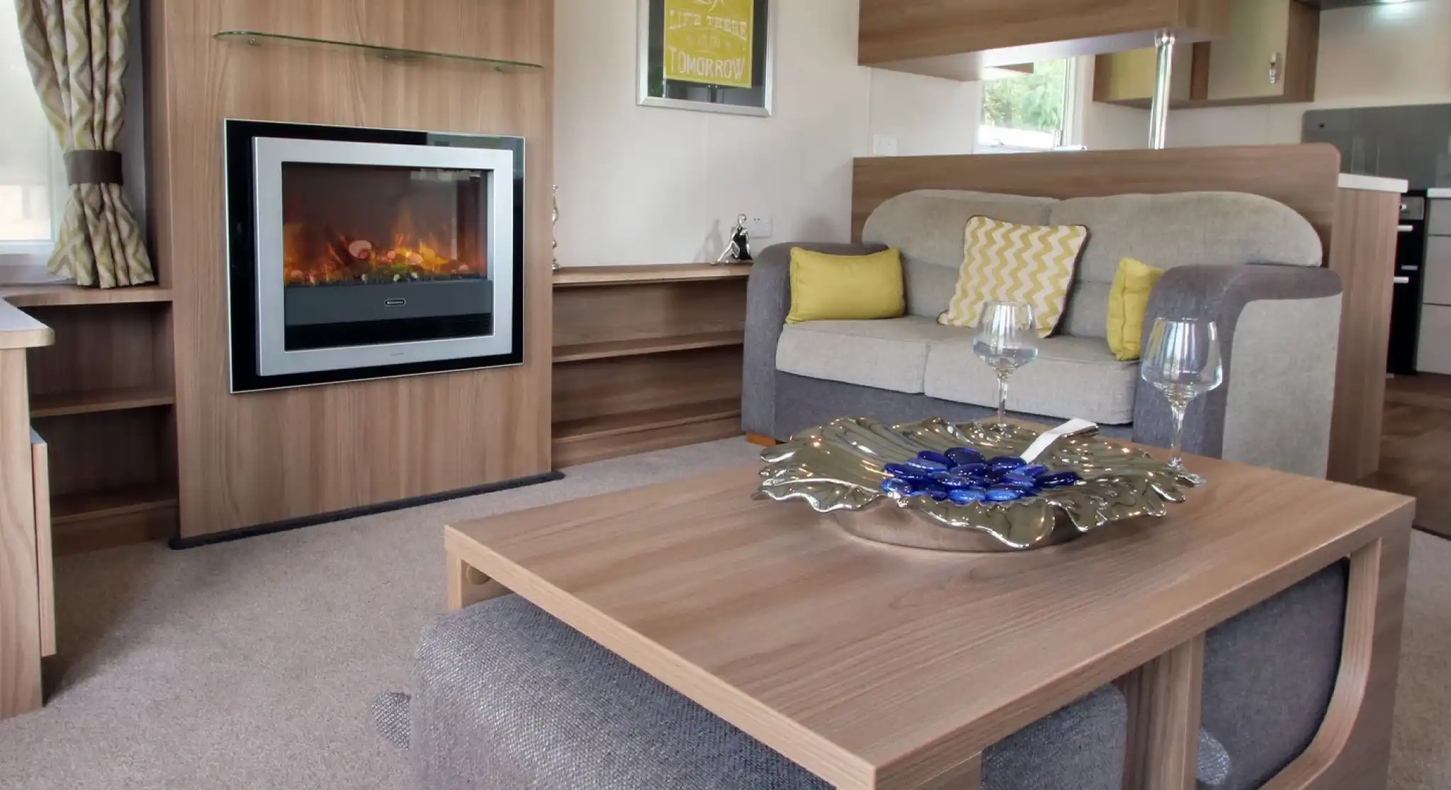 A cozy living area featuring a comfortable gray and beige sofa with decorative cushions, a modern electric fireplace, and a stylish coffee table with a decorative bowl. The room is well-lit by natural light, with curtains and wall art complementing the warm ambiance.