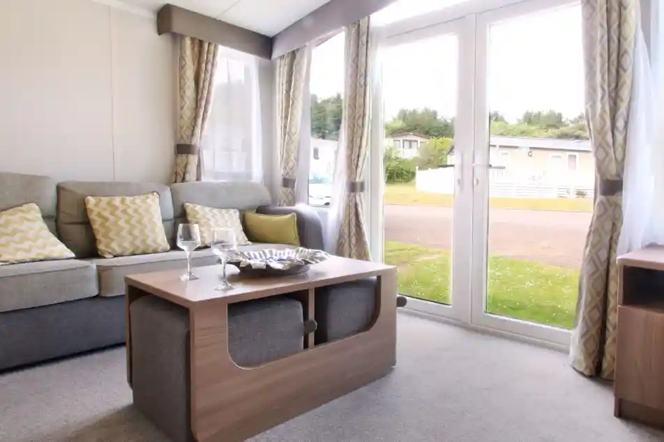 A cozy living area featuring a grey sofa with yellow patterned cushions, a wooden coffee table with a decorative bowl, and large glass doors opening to the outdoors. Soft curtains frame the windows, and the space has a warm, inviting ambiance.