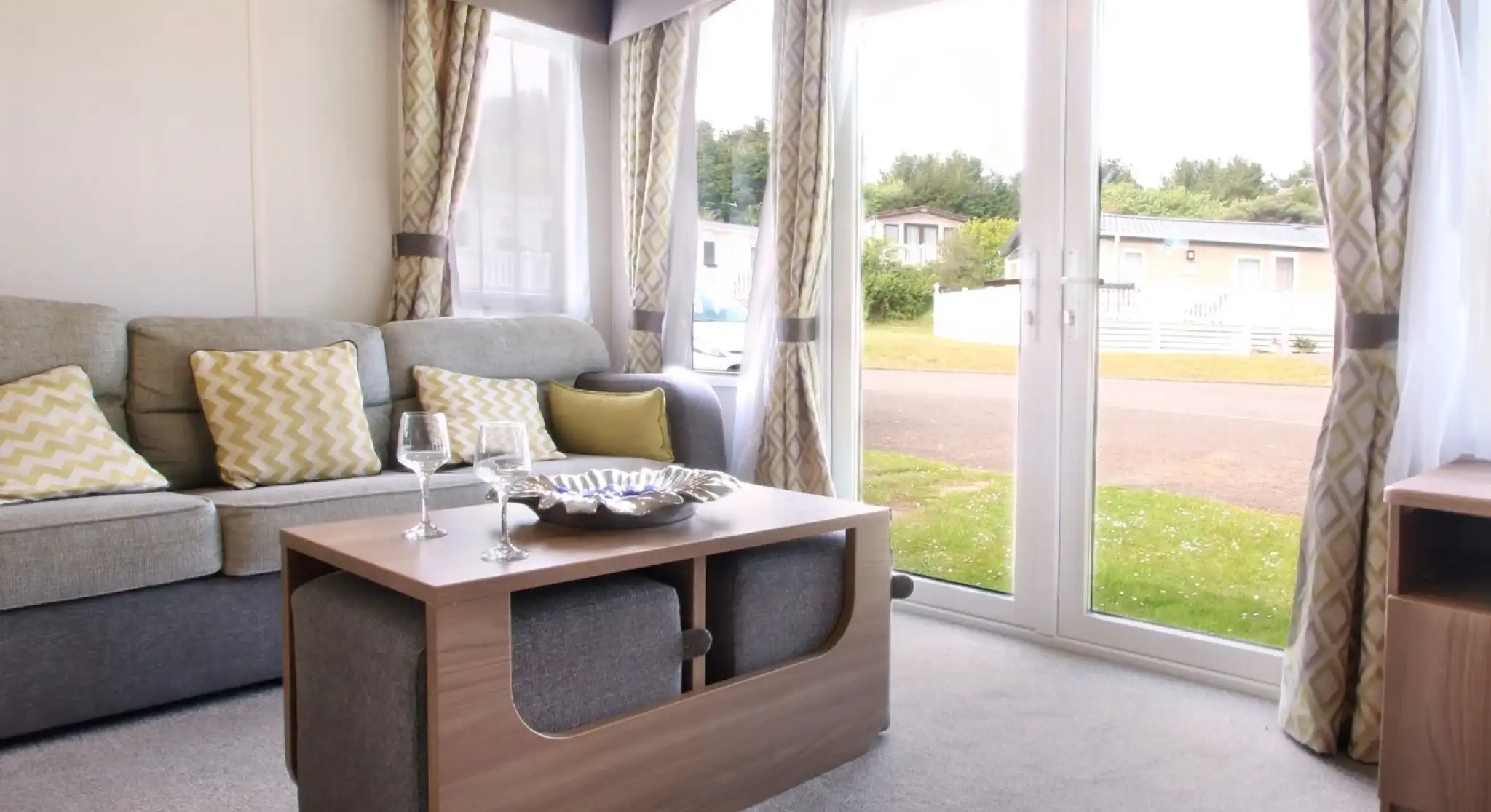 A cozy living area featuring a grey sofa with yellow patterned cushions, a wooden coffee table with a decorative bowl, and large glass doors opening to the outdoors. Soft curtains frame the windows, and the space has a warm, inviting ambiance.