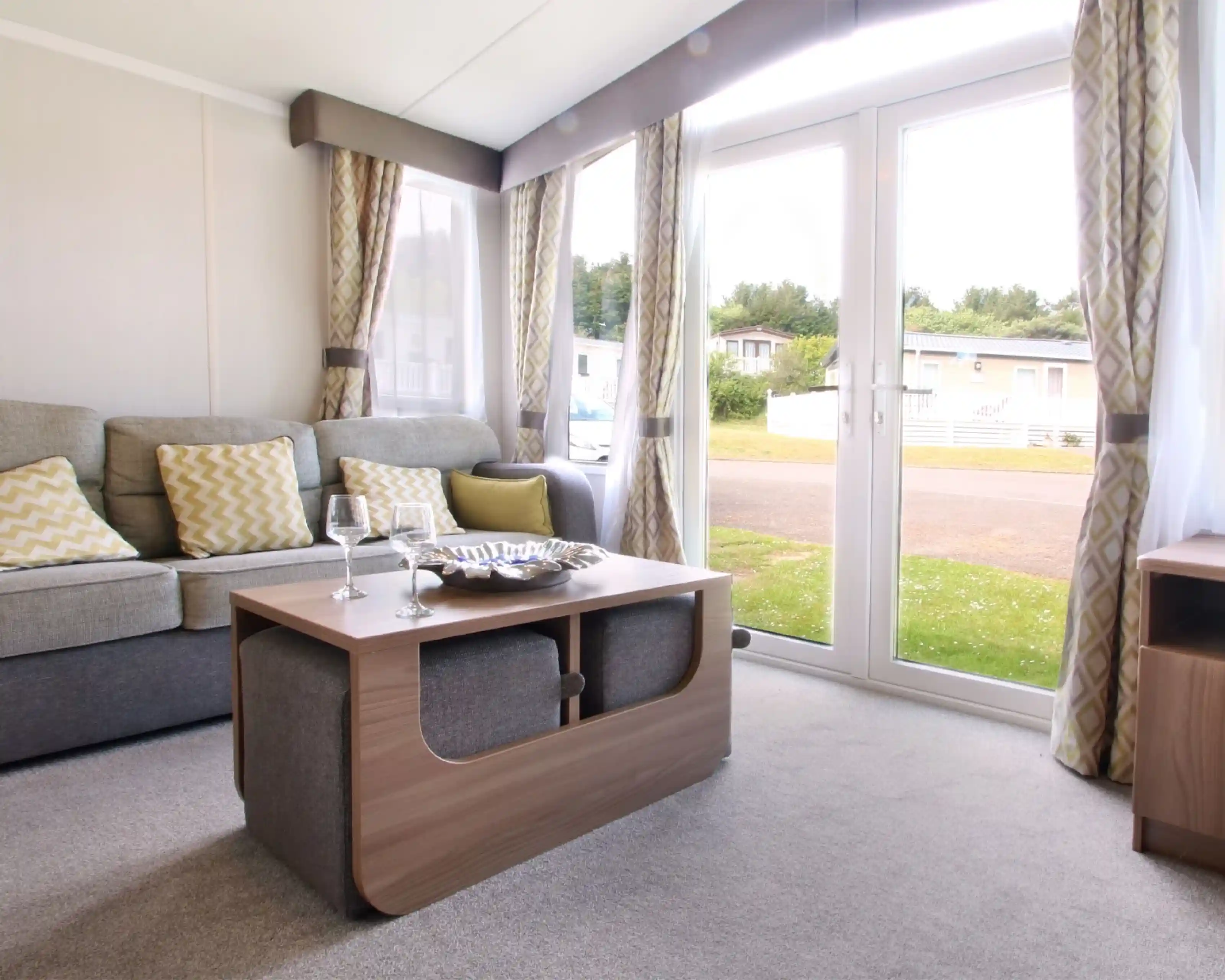 A cozy living area featuring a grey sofa with yellow patterned cushions, a wooden coffee table with a decorative bowl, and large glass doors opening to the outdoors. Soft curtains frame the windows, and the space has a warm, inviting ambiance.