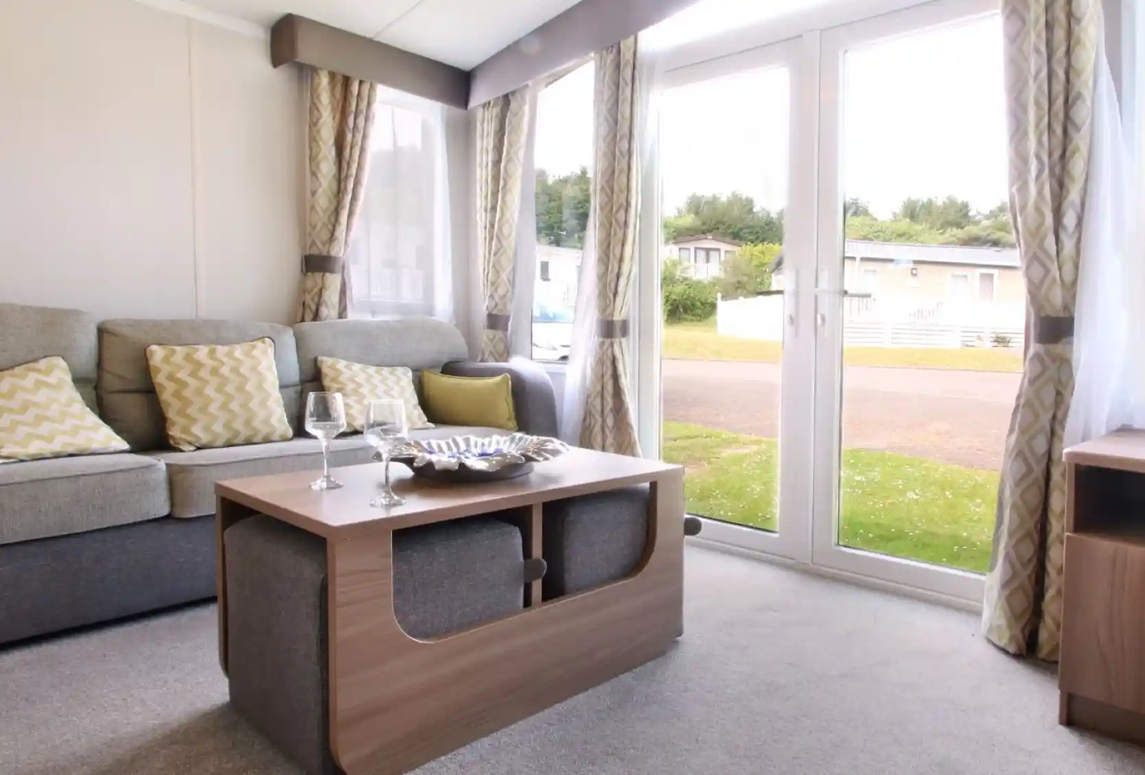A cozy living area featuring a grey sofa with yellow patterned cushions, a wooden coffee table with a decorative bowl, and large glass doors opening to the outdoors. Soft curtains frame the windows, and the space has a warm, inviting ambiance.