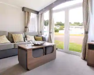 A cozy living area featuring a grey sofa with yellow patterned cushions, a wooden coffee table with a decorative bowl, and large glass doors opening to the outdoors. Soft curtains frame the windows, and the space has a warm, inviting ambiance.