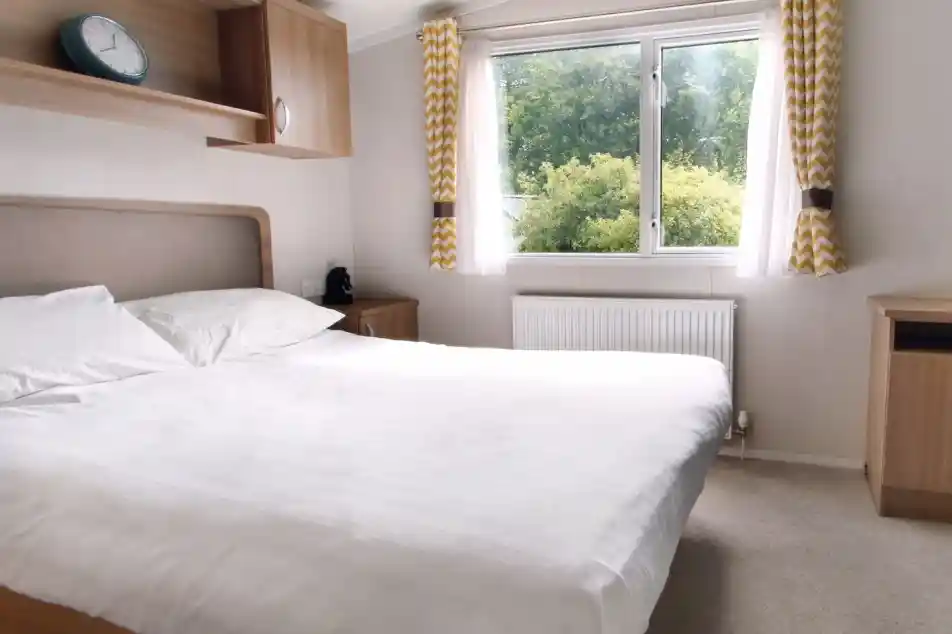 A cozy bedroom featuring a large bed with white linens, a wooden bedside table, and a clock above the headboard. Natural light streams in through a window adorned with yellow and white patterned curtains, illuminating a view of greenery outside. A small radiator sits beneath the window.