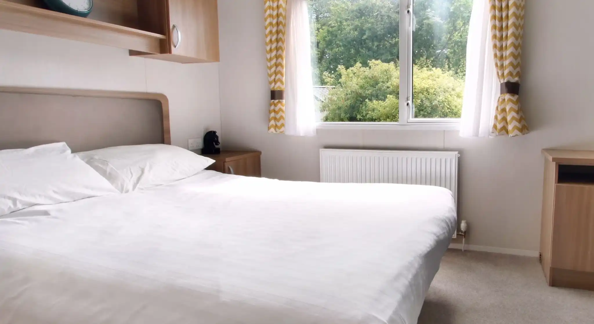 A cozy bedroom featuring a large bed with white linens, a wooden bedside table, and a clock above the headboard. Natural light streams in through a window adorned with yellow and white patterned curtains, illuminating a view of greenery outside. A small radiator sits beneath the window.