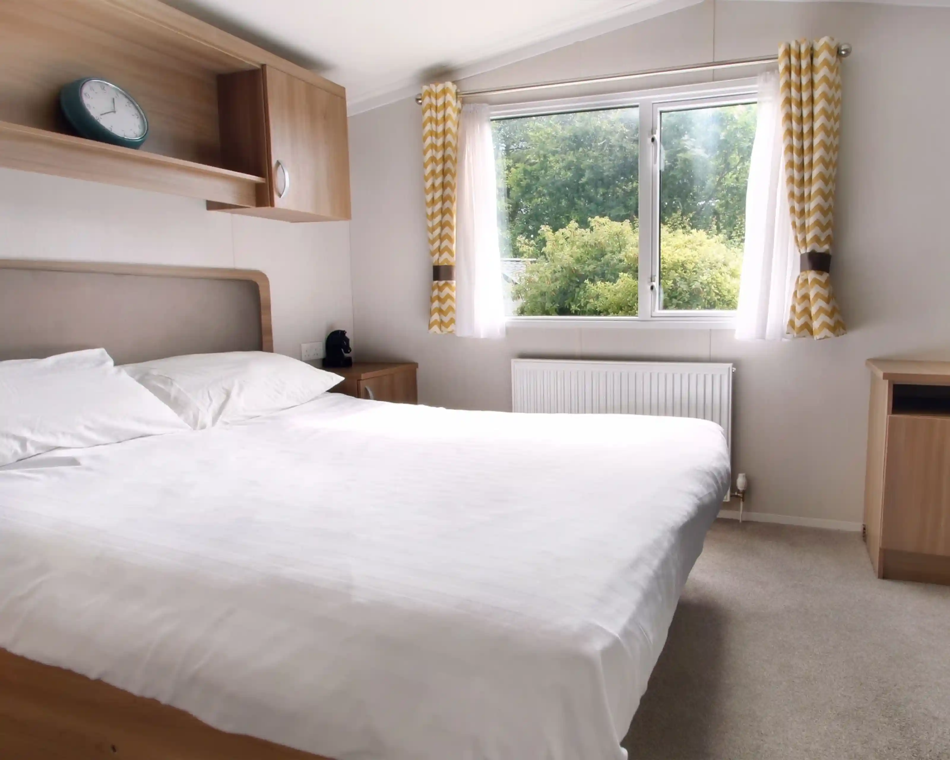 A cozy bedroom featuring a large bed with white linens, a wooden bedside table, and a clock above the headboard. Natural light streams in through a window adorned with yellow and white patterned curtains, illuminating a view of greenery outside. A small radiator sits beneath the window.
