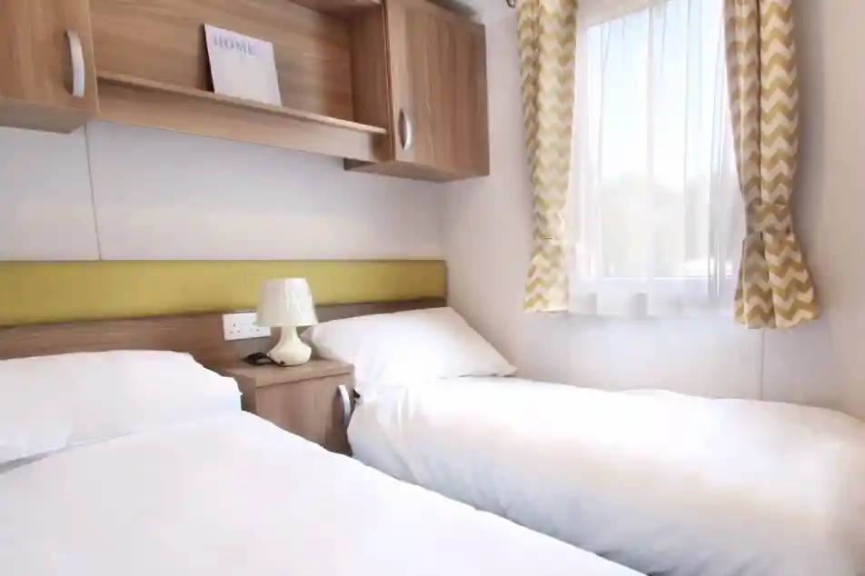 A cozy bedroom featuring two single beds with white bedding, a wooden nightstand with a lamp, and a modern window draped with chevron-patterned curtains. A decorative sign reading "HOME" is visible on a shelf above the beds.