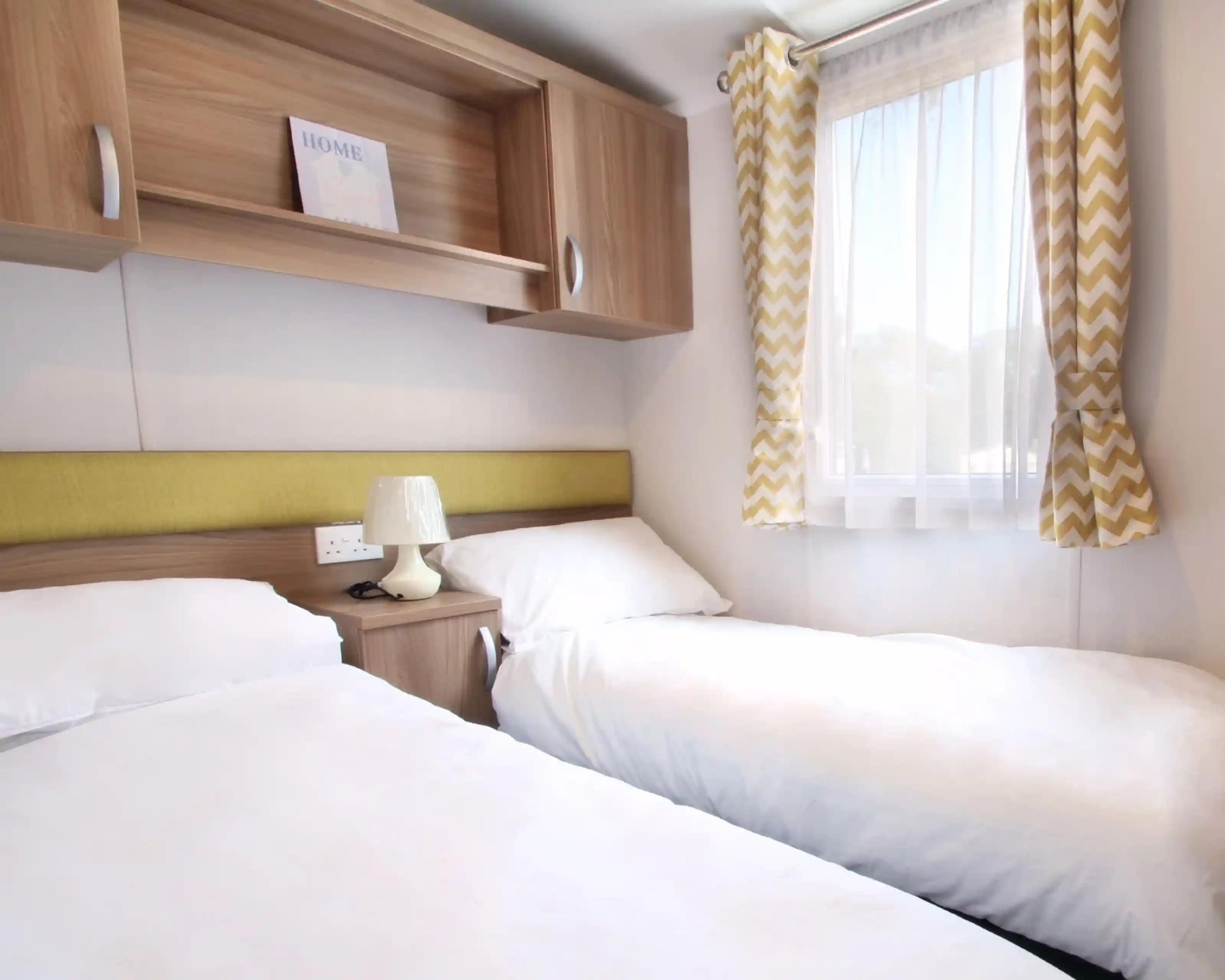 A cozy bedroom featuring two single beds with white bedding, a wooden nightstand with a lamp, and a modern window draped with chevron-patterned curtains. A decorative sign reading "HOME" is visible on a shelf above the beds.