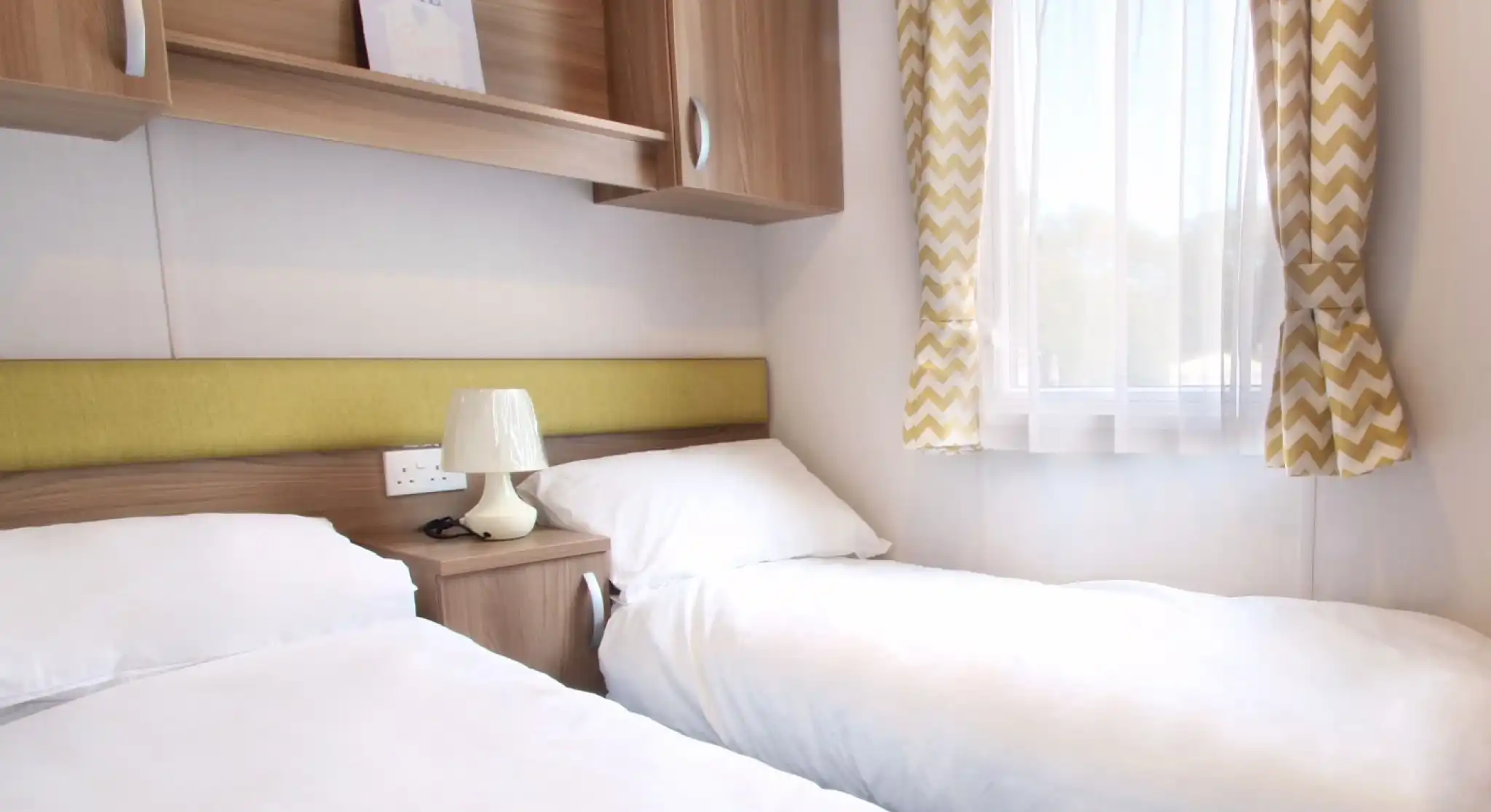 A cozy bedroom featuring two single beds with white bedding, a wooden nightstand with a lamp, and a modern window draped with chevron-patterned curtains. A decorative sign reading "HOME" is visible on a shelf above the beds.