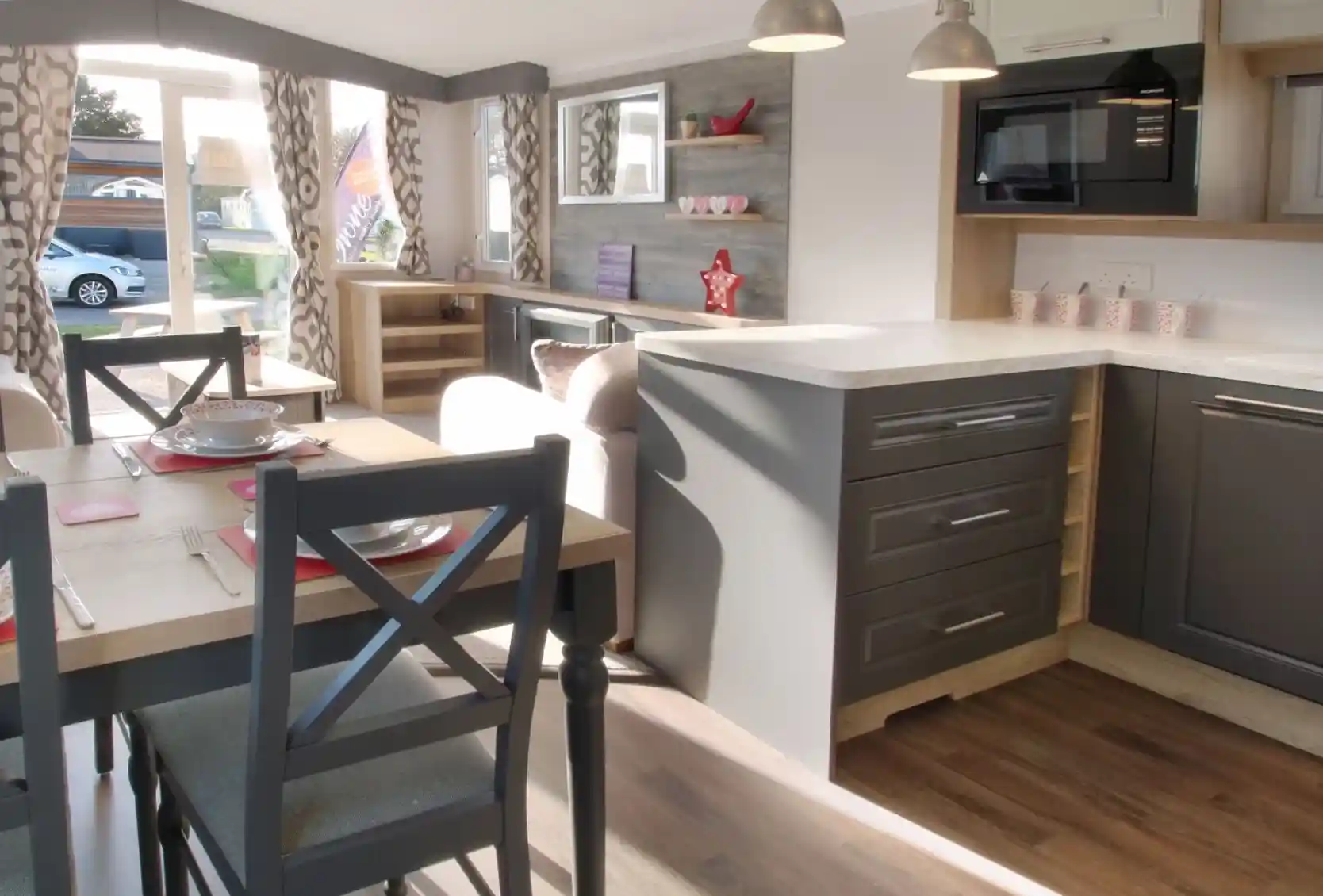 A modern interior of a caravan featuring a kitchen with gray cabinets, a white countertop, and a microwave. A dining table with gray chairs is set for a meal, and large windows allow natural light to brighten the space. Decor includes curtains with a geometric pattern and decorative items on shelves.