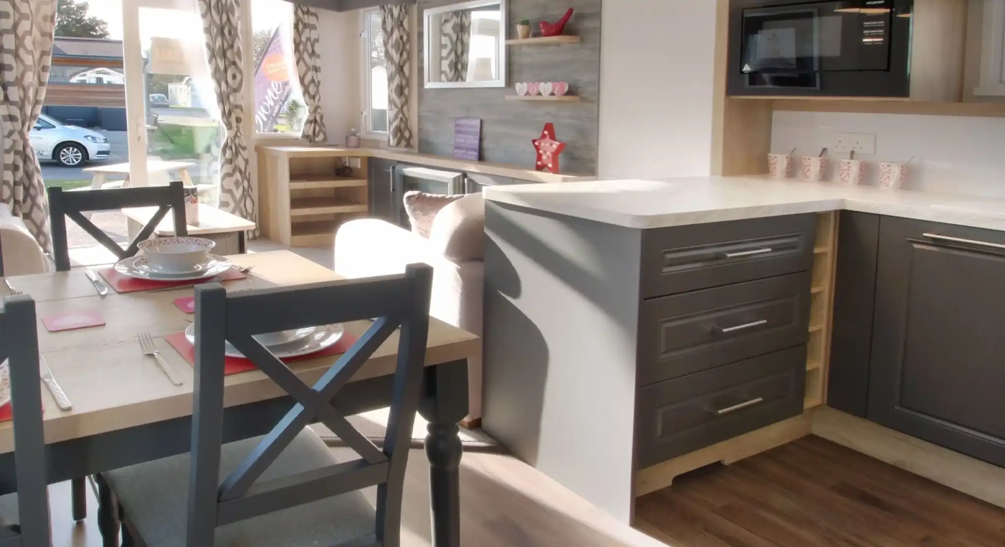 A modern interior of a caravan featuring a kitchen with gray cabinets, a white countertop, and a microwave. A dining table with gray chairs is set for a meal, and large windows allow natural light to brighten the space. Decor includes curtains with a geometric pattern and decorative items on shelves.