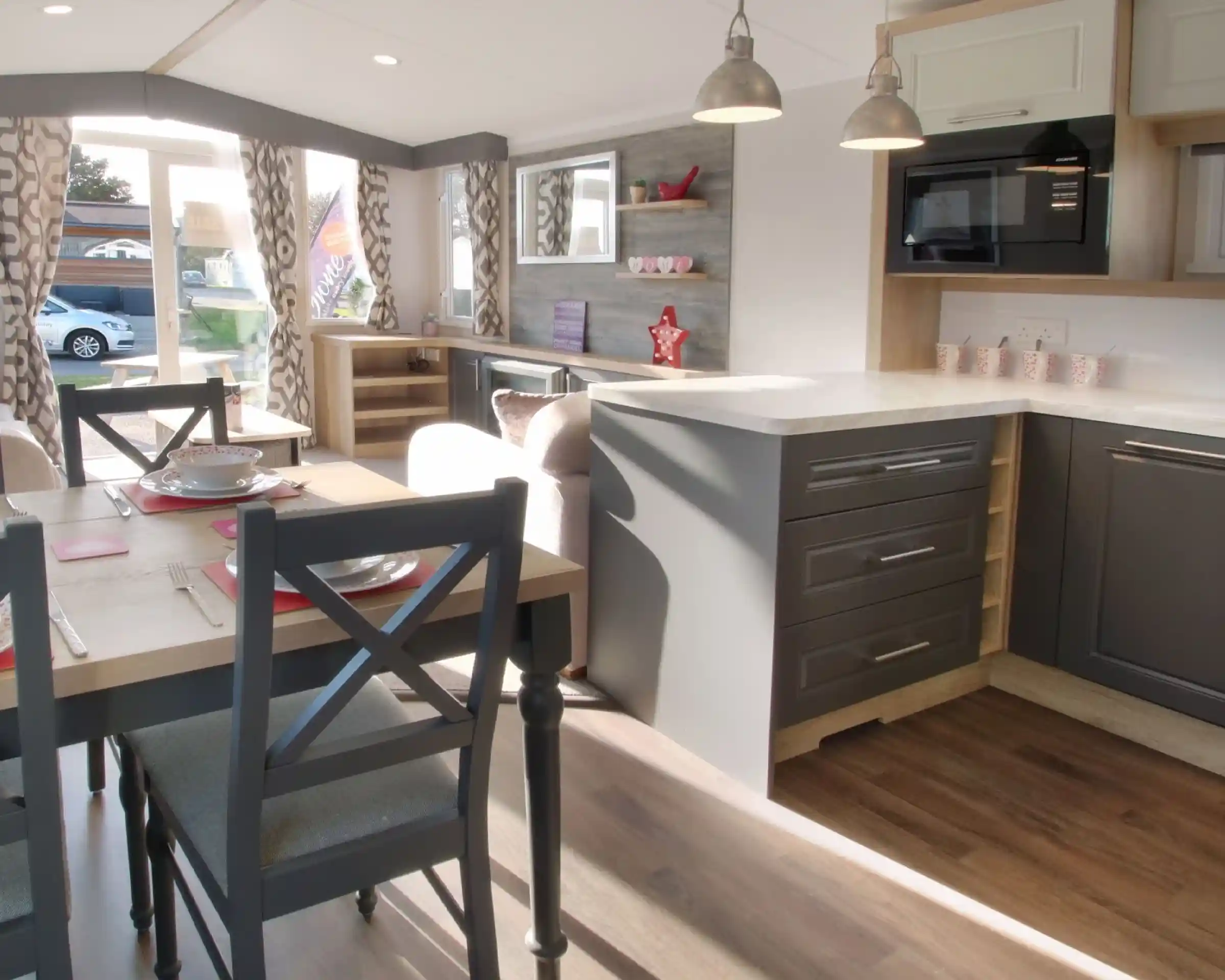 A modern interior of a caravan featuring a kitchen with gray cabinets, a white countertop, and a microwave. A dining table with gray chairs is set for a meal, and large windows allow natural light to brighten the space. Decor includes curtains with a geometric pattern and decorative items on shelves.