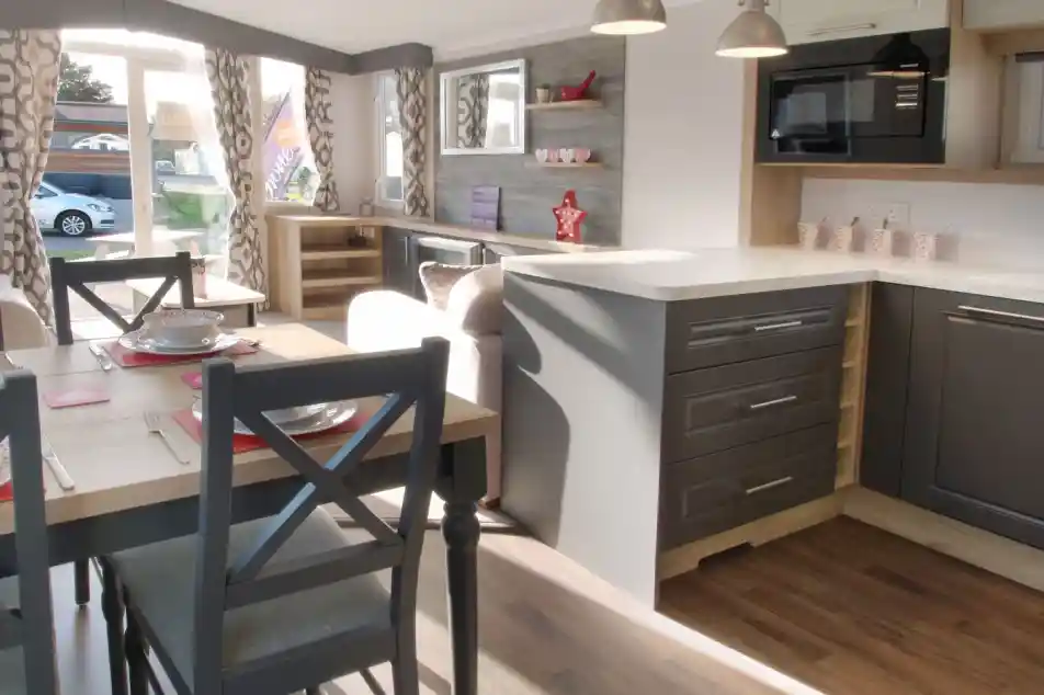 A modern interior of a caravan featuring a kitchen with gray cabinets, a white countertop, and a microwave. A dining table with gray chairs is set for a meal, and large windows allow natural light to brighten the space. Decor includes curtains with a geometric pattern and decorative items on shelves.