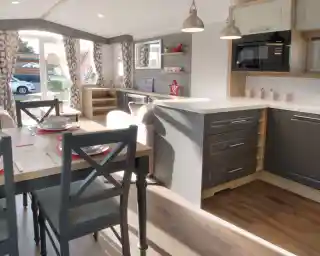 A modern interior of a caravan featuring a kitchen with gray cabinets, a white countertop, and a microwave. A dining table with gray chairs is set for a meal, and large windows allow natural light to brighten the space. Decor includes curtains with a geometric pattern and decorative items on shelves.