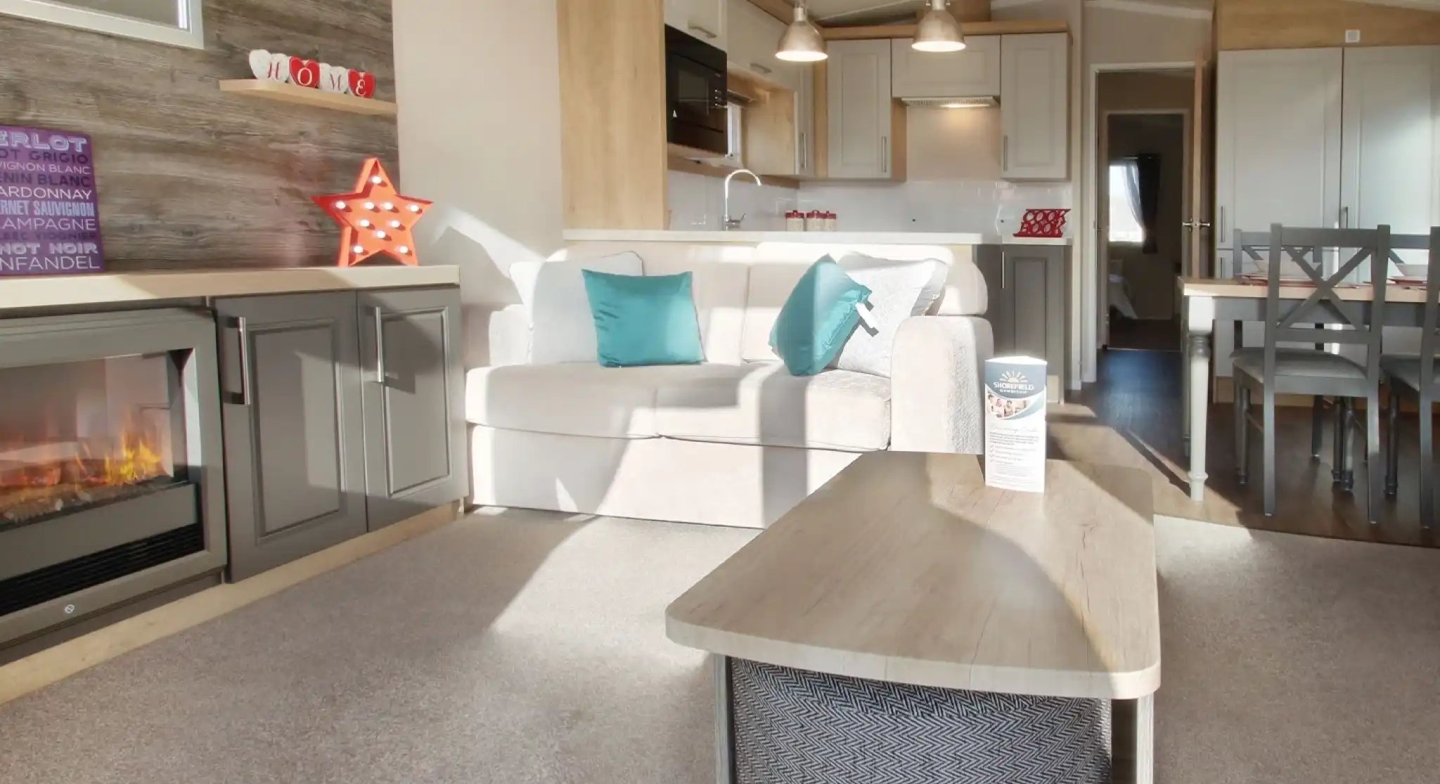 A cozy living area features a light-colored sofa with teal cushions, a wooden coffee table, and a modern electric fireplace. The kitchen, visible in the background, has white cabinets and pendant lighting, complemented by a dining area with a wooden table and chairs. A decorative star and various items adorn the shelves. Natural light brightens the space.