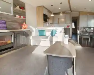 A cozy living area features a light-colored sofa with teal cushions, a wooden coffee table, and a modern electric fireplace. The kitchen, visible in the background, has white cabinets and pendant lighting, complemented by a dining area with a wooden table and chairs. A decorative star and various items adorn the shelves. Natural light brightens the space.