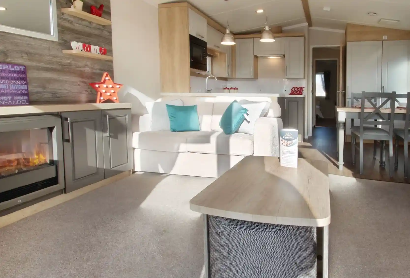 A cozy living area features a light-colored sofa with teal cushions, a wooden coffee table, and a modern electric fireplace. The kitchen, visible in the background, has white cabinets and pendant lighting, complemented by a dining area with a wooden table and chairs. A decorative star and various items adorn the shelves. Natural light brightens the space.