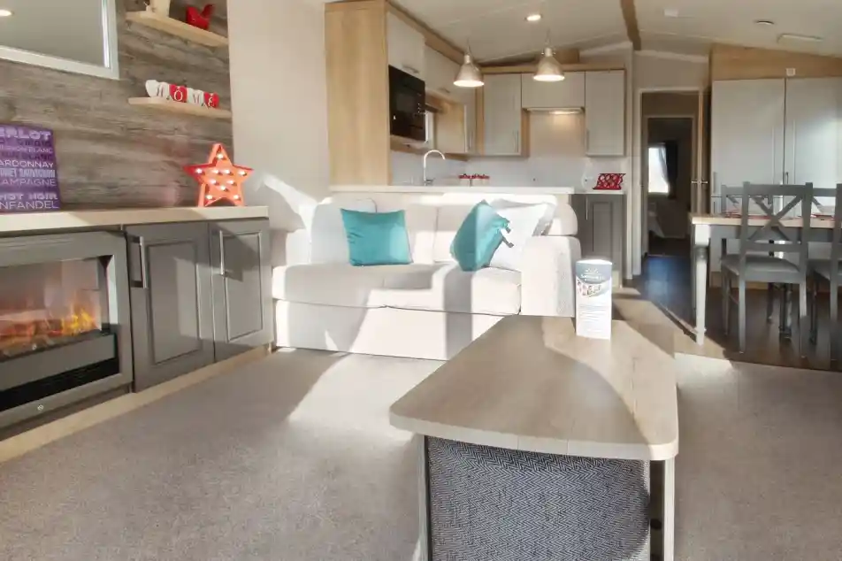 A cozy living area features a light-colored sofa with teal cushions, a wooden coffee table, and a modern electric fireplace. The kitchen, visible in the background, has white cabinets and pendant lighting, complemented by a dining area with a wooden table and chairs. A decorative star and various items adorn the shelves. Natural light brightens the space.