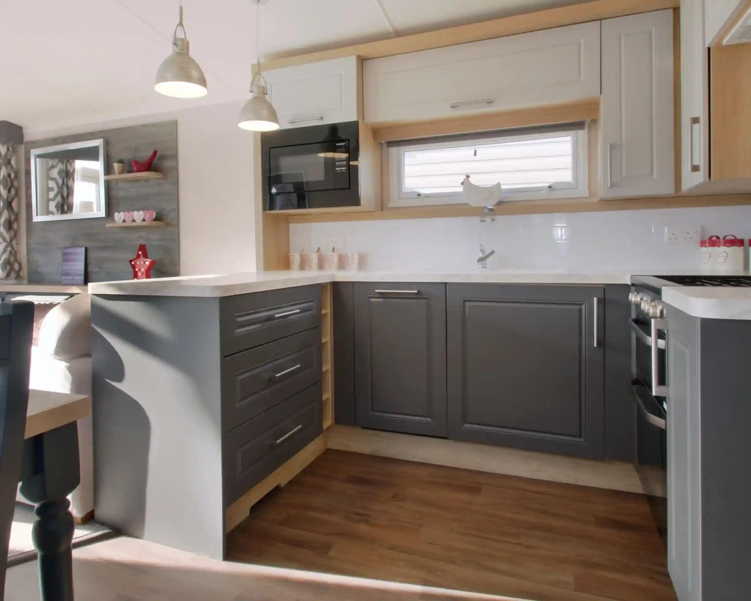 A modern kitchen featuring gray and white cabinetry, a countertop with a glossy finish, and silver pendant lights. A microwave is mounted above the counter, and wooden flooring extends throughout the space. Shelves in the background display decorative items.