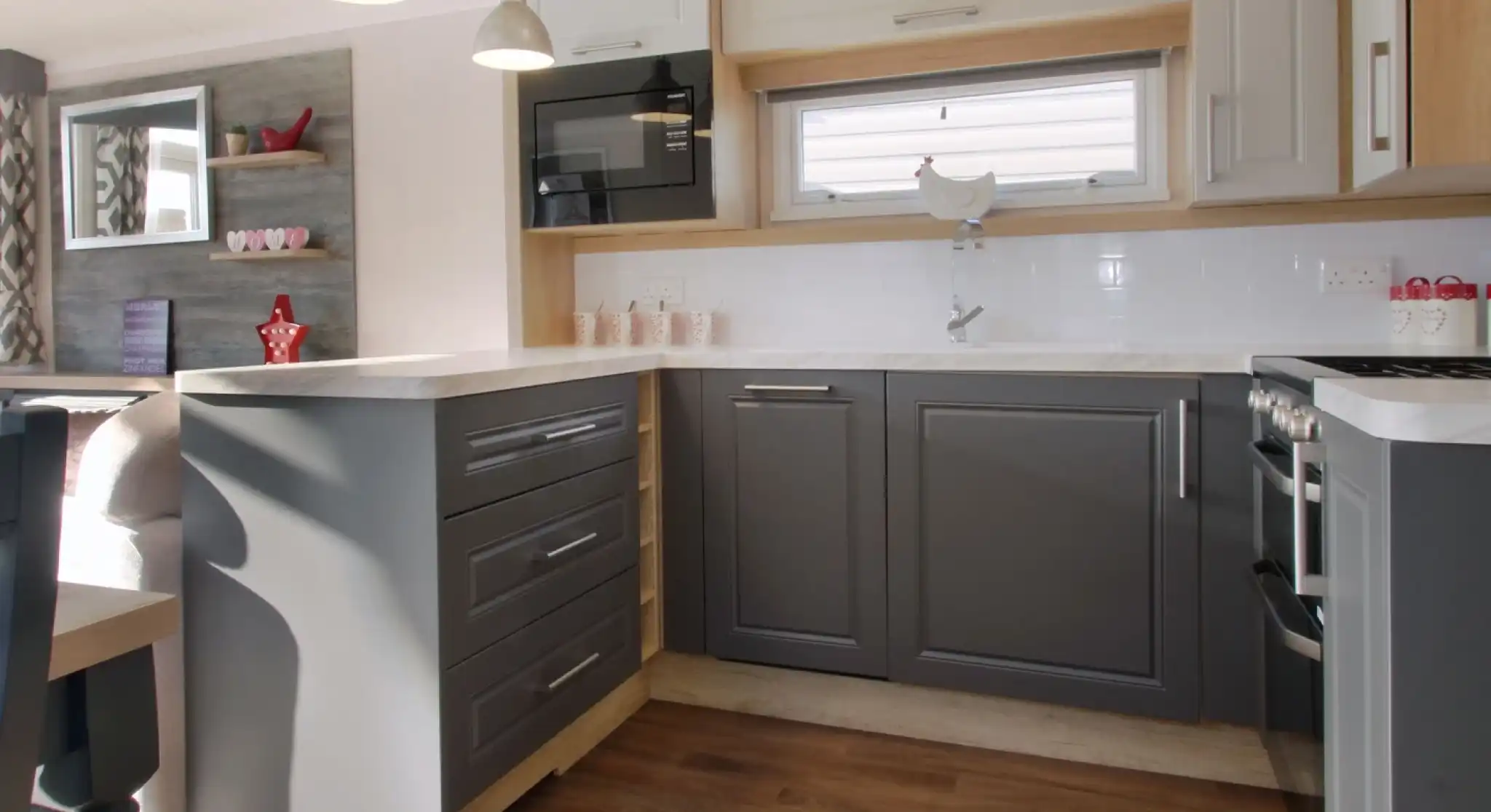A modern kitchen featuring gray and white cabinetry, a countertop with a glossy finish, and silver pendant lights. A microwave is mounted above the counter, and wooden flooring extends throughout the space. Shelves in the background display decorative items.