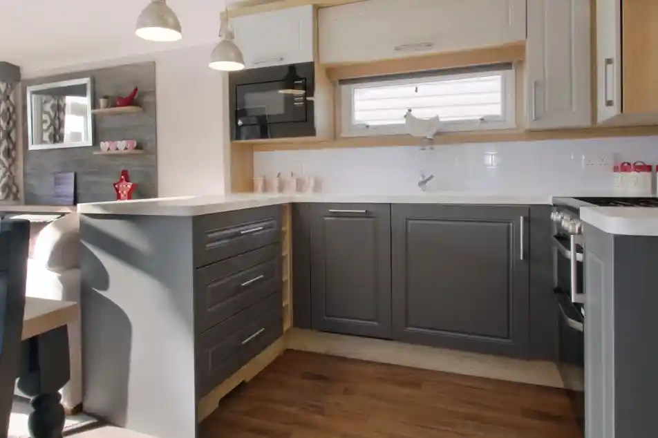 A modern kitchen featuring gray and white cabinetry, a countertop with a glossy finish, and silver pendant lights. A microwave is mounted above the counter, and wooden flooring extends throughout the space. Shelves in the background display decorative items.